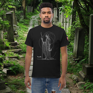 Man wearing a black Grim Sentinel t-shirt with white line art of a cloaked reaper holding a scythe and skull, standing on a mossy cemetery path.