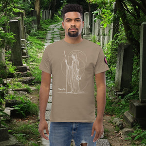 Man wearing a brown Savana Grim Sentinel t-shirt with white line art of a cloaked reaper holding a scythe and skull, standing on a mossy cemetery path.
