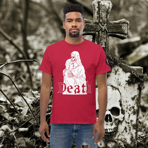 Man wearing a cardinal red Death’s Embrace t-shirt featuring skeleton holding a woman, gothic lettering reading “Death,” standing before a weathered grave.