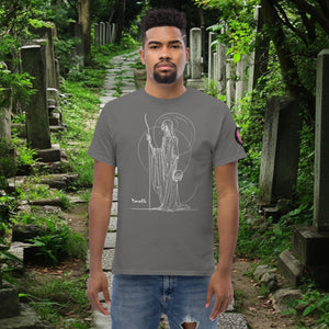 Man wearing a charcoal Grim Sentinel t-shirt with white line art of a cloaked reaper holding a scythe and skull, standing on a mossy cemetery path.