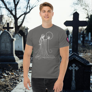 Man wearing a charcoal Eternal Vigil t-shirt with white line art of a robed figure holding a skull, standing in a cemetery surrounded by tombstones.