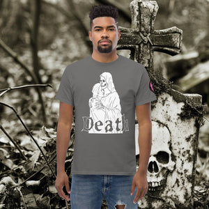 Man wearing a charcoal Death’s Embrace t-shirt featuring skeleton holding a woman, gothic lettering reading “Death,” standing before a weathered grave.