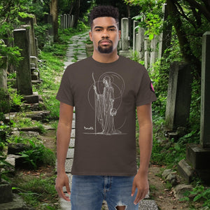 Man wearing a dark chocolate Grim Sentinel t-shirt with white line art of a cloaked reaper holding a scythe and skull, standing on a mossy cemetery path.