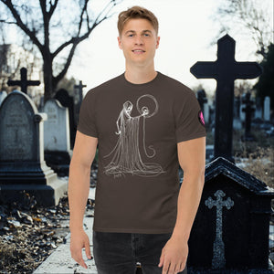 Man wearing a dark chocolate Eternal Vigil t-shirt with white line art of a robed figure holding a skull, standing in a cemetery surrounded by tombstones.
