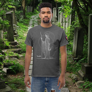 Man wearing a dark heather Grim Sentinel t-shirt with white line art of a cloaked reaper holding a scythe and skull, standing on a mossy cemetery path.