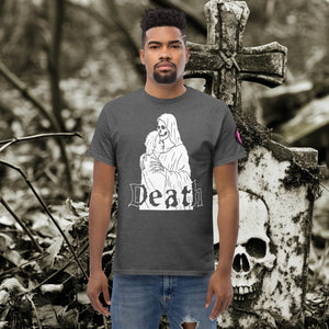 Man wearing a dark heather Death’s Embrace t-shirt featuring skeleton holding a woman, gothic lettering reading “Death,” standing before a weathered grave.