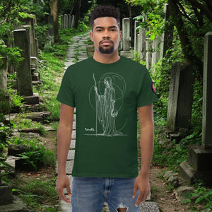 Man wearing a forest green Grim Sentinel t-shirt with white line art of a cloaked reaper holding a scythe and skull, standing on a mossy cemetery path.