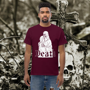 Man wearing a maroon Death’s Embrace t-shirt featuring skeleton holding a woman, gothic lettering reading “Death,” standing before a weathered grave.