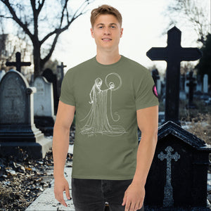 Man wearing a military green Eternal Vigil t-shirt with white line art of a robed figure holding a skull, standing in a cemetery surrounded by tombstones.