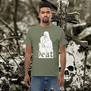 Man wearing a military green Death’s Embrace t-shirt featuring skeleton holding a woman, gothic lettering reading “Death,” standing before a weathered grave.