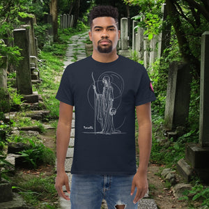 Man wearing a navy Grim Sentinel t-shirt with white line art of a cloaked reaper holding a scythe and skull, standing on a mossy cemetery path.