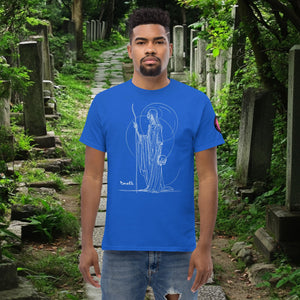 Man wearing a royal blue Grim Sentinel t-shirt with white line art of a cloaked reaper holding a scythe and skull, standing on a mossy cemetery path.