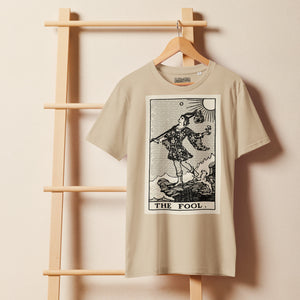 Mid-light organic cotton t-shirt featuring The Fool tarot card with cliff, dog, and sun in high-contrast woodcut rendering.