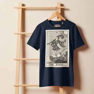 Mid-light organic cotton t-shirt featuring The Fool tarot card with cliff, dog, and sun in high-contrast woodcut rendering.