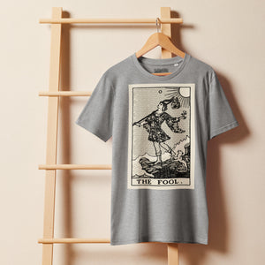 Mid-light organic cotton t-shirt featuring The Fool tarot card with cliff, dog, and sun in high-contrast woodcut rendering.