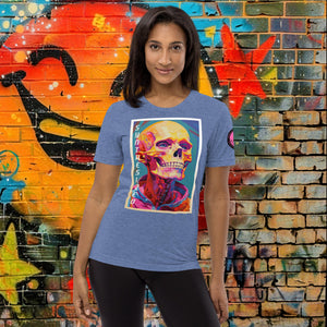 A woman wearing a blue triblend Synthesized Futility Premium Tee with vibrant stylized skull, graffiti background with orange and blue tones.