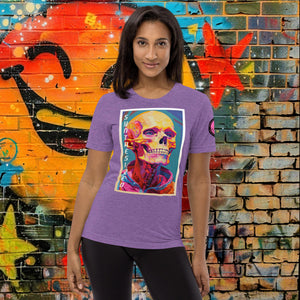 A woman wearing a purple triblend Synthesized Futility Premium Tee with vibrant stylized skull, graffiti background with orange and blue tones.