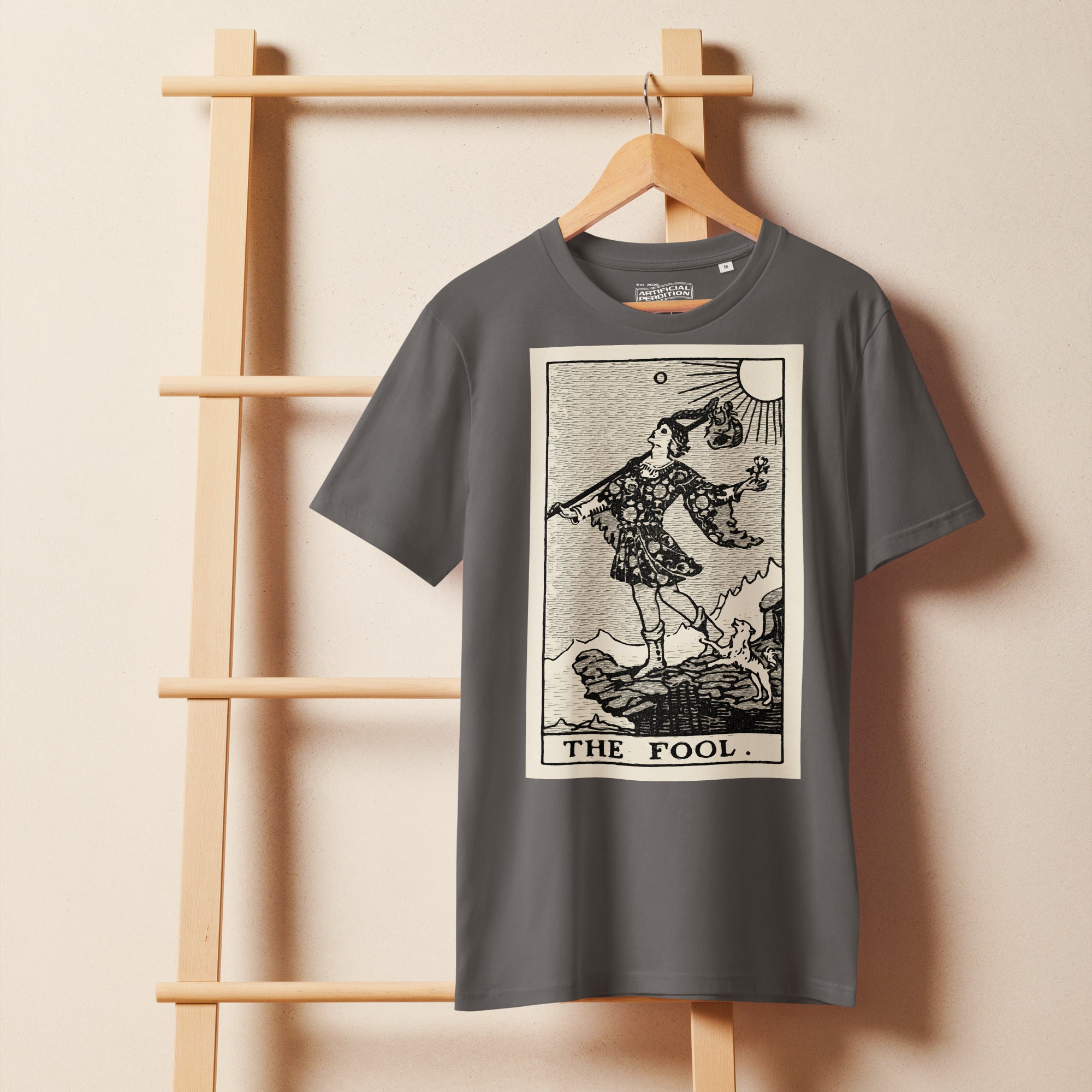 Mid-light organic cotton t-shirt featuring The Fool tarot card with cliff, dog, and sun in high-contrast woodcut rendering.