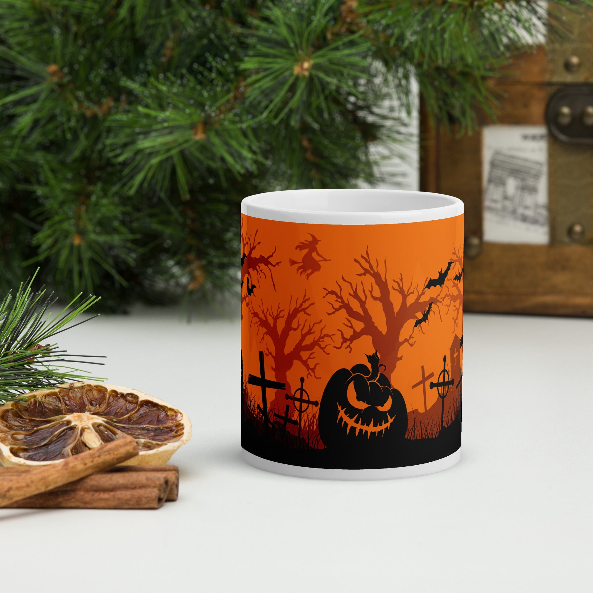 White glossy ceramic mug featuring a wrap-around graveyard illustration with black silhouettes of tombstones, trees, bats, and a jack-o’-lantern against an orange background.