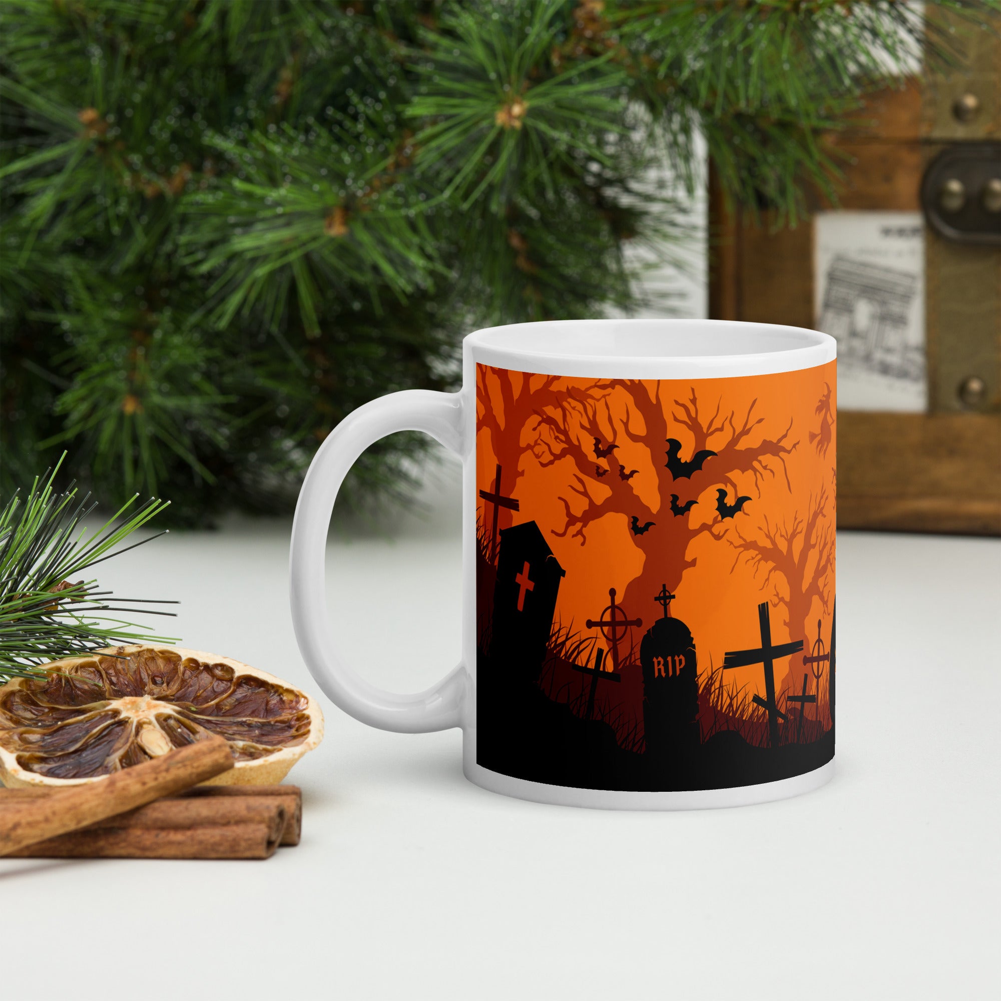 White glossy ceramic mug featuring a wrap-around graveyard illustration with black silhouettes of tombstones, trees, bats, and a jack-o’-lantern against an orange background.